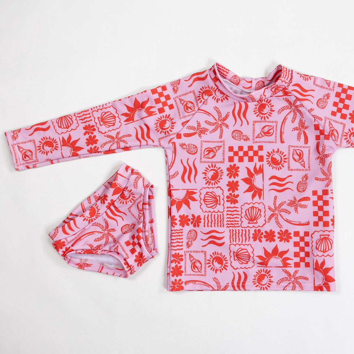 A children's long-sleeve swim top and matching swim bottoms laid flat on a white background. The swimwear features a pink base with a red tropical and beach-themed pattern of palm trees, suns, waves, and geometric shapes.