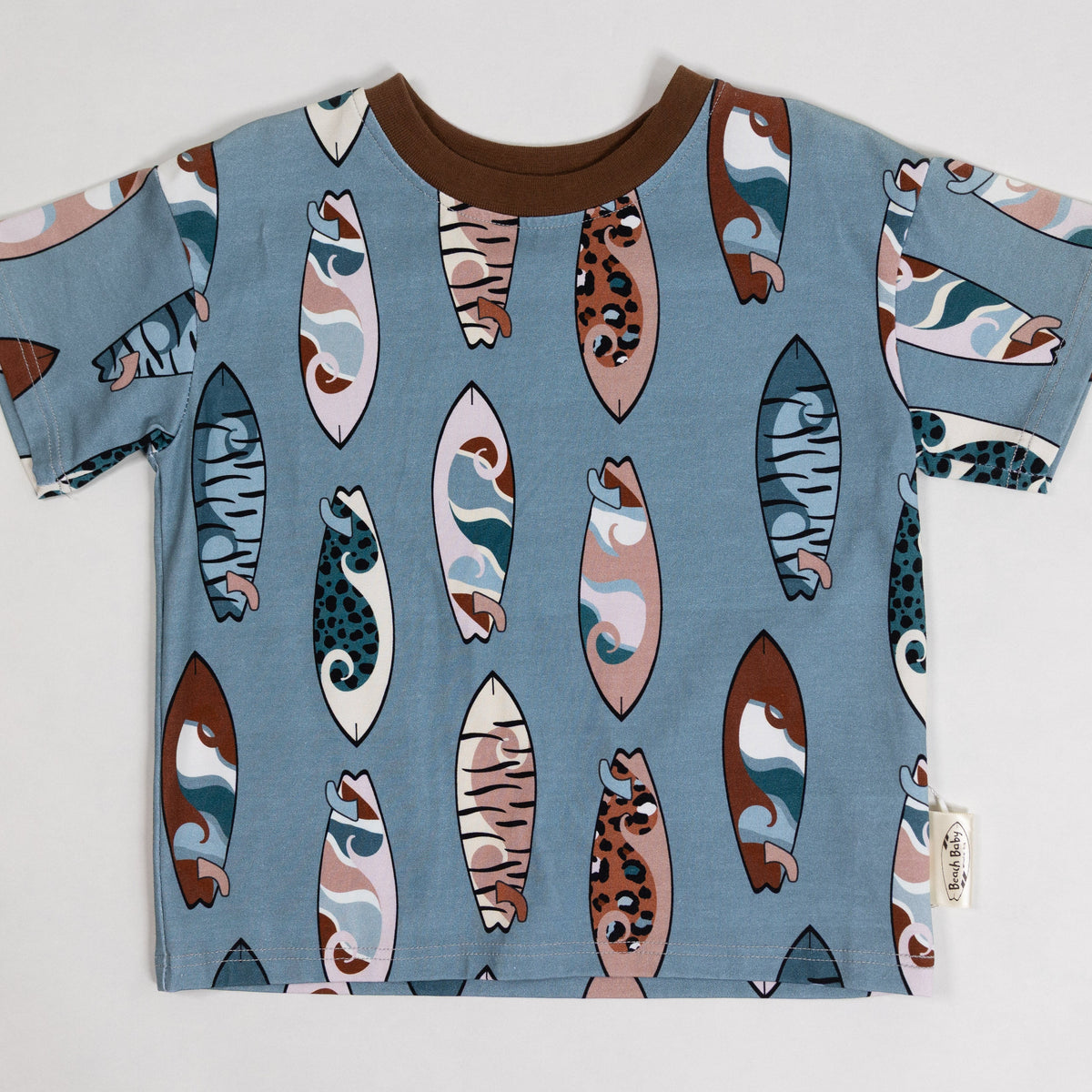 A short-sleeve kids' t-shirt in dusty blue, featuring an all-over surfboard pattern in brown, beige, and white, with a solid brown crew neck.
