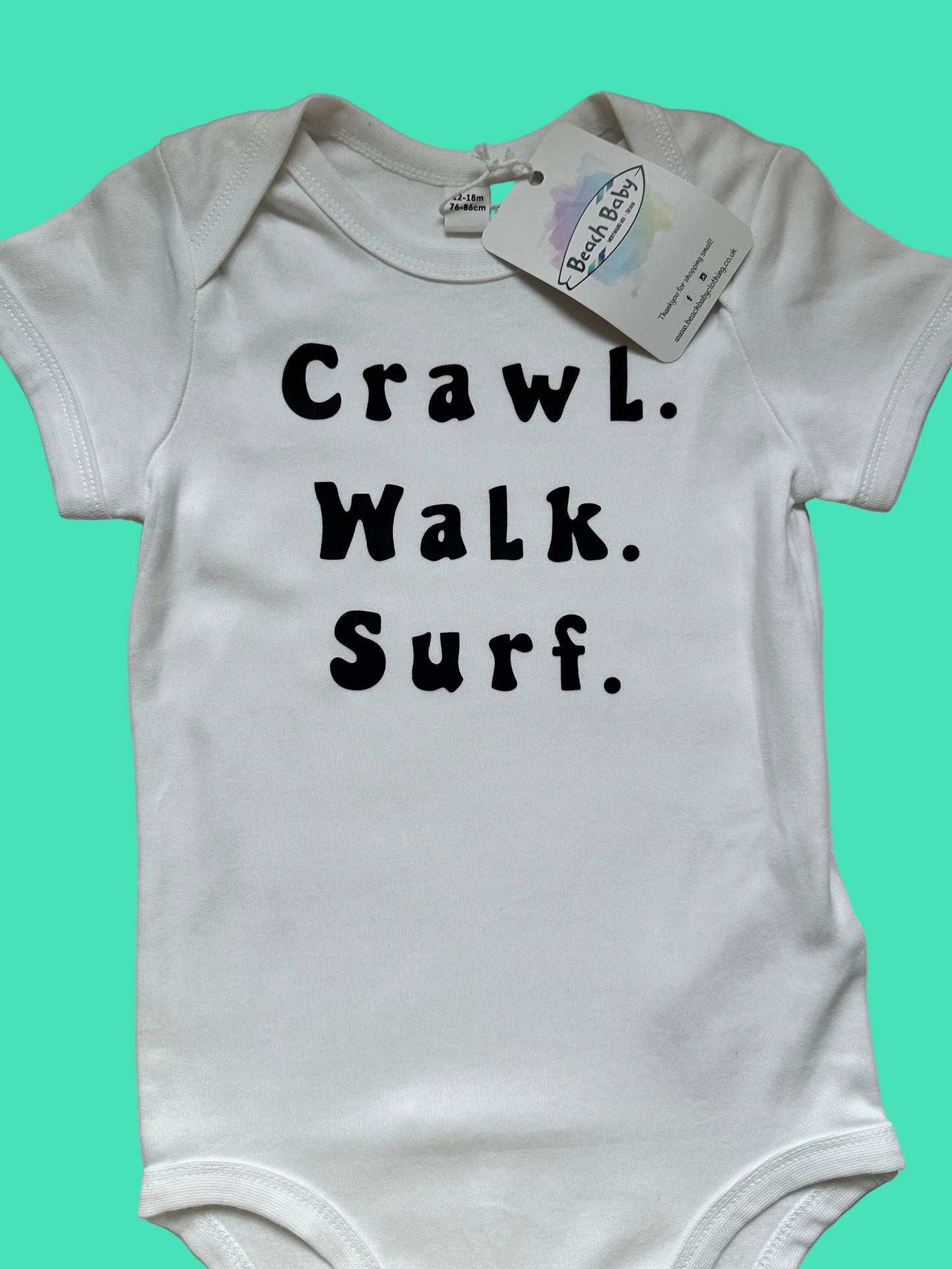 White baby onesie with text 'Crawl. Walk. Surf.' and a tag on a light gray background