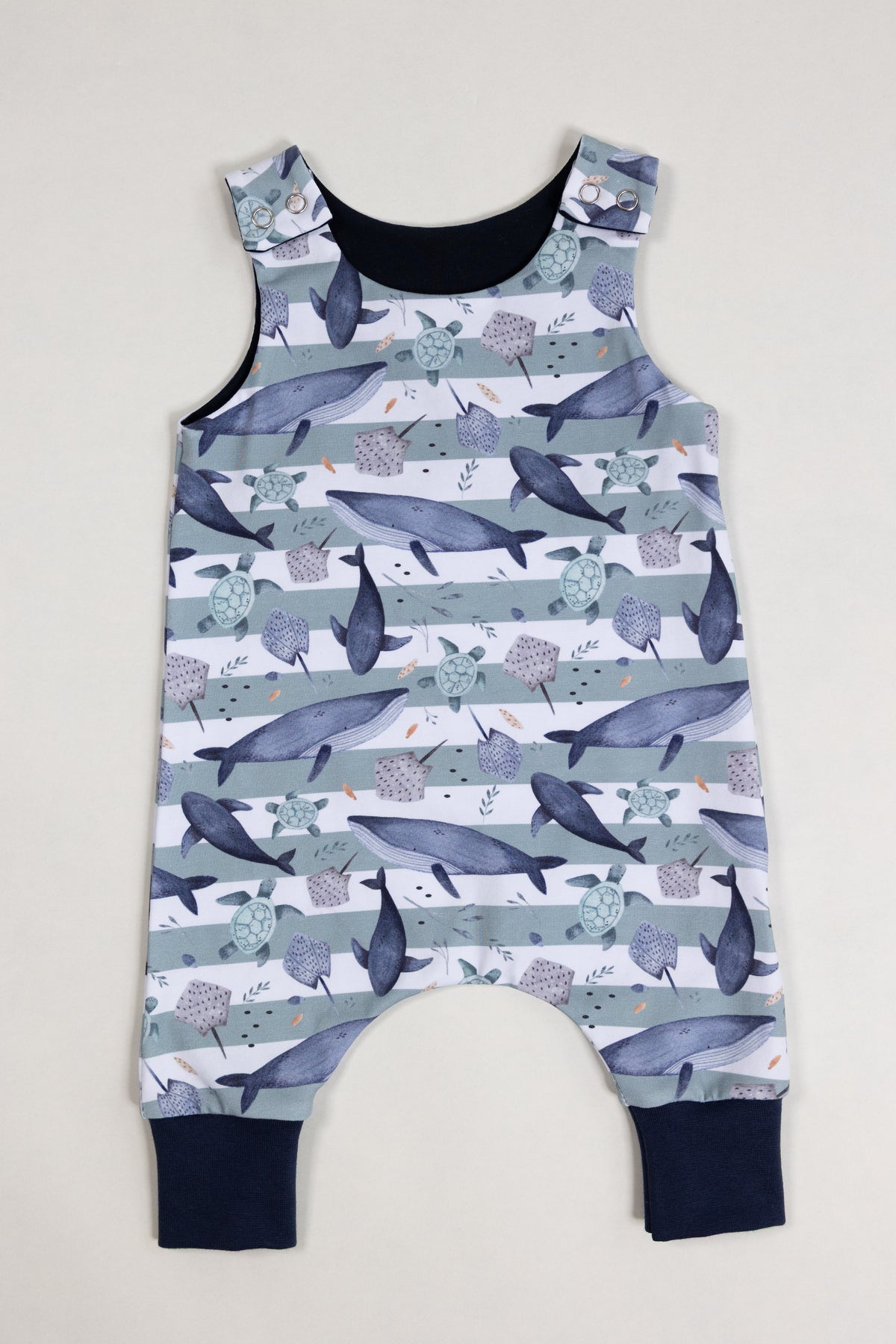 A sleeveless baby romper with an all-over whale print, featuring dark navy blue trim at the neckline and leg cuffs, laid flat against a light beige background.
