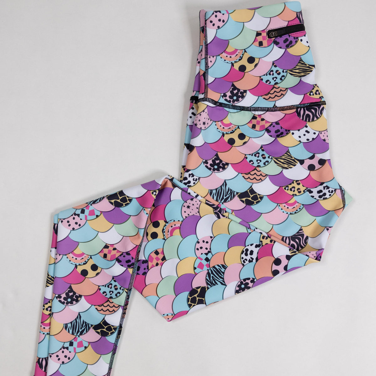 A pair of folded leggings with a colorful mermaid scale pattern featuring pink, purple, blue, yellow, and black-and-white spots, laid flat on a light gray background.