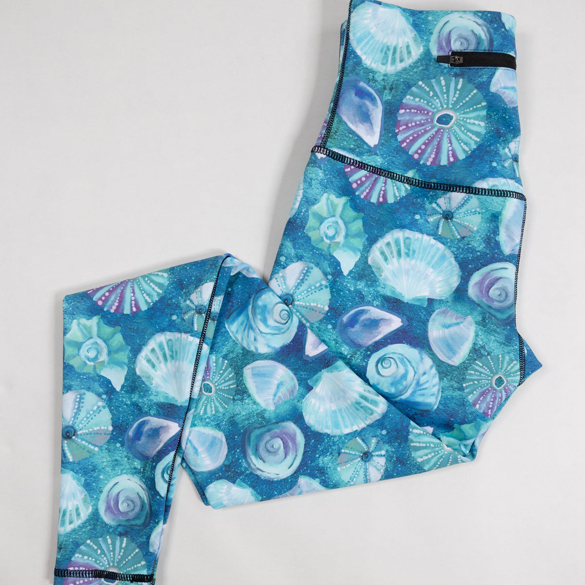 A pair of high-waisted leggings with a vibrant blue and purple seashell pattern, laid flat on a light gray background.