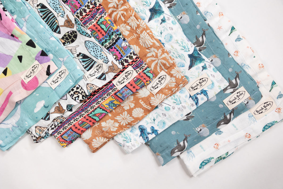 A fan arrangement of colorful reusable cloth muslins with various printed patterns including sea life, tropical leaves, and abstract designs, displayed on a clean white background.