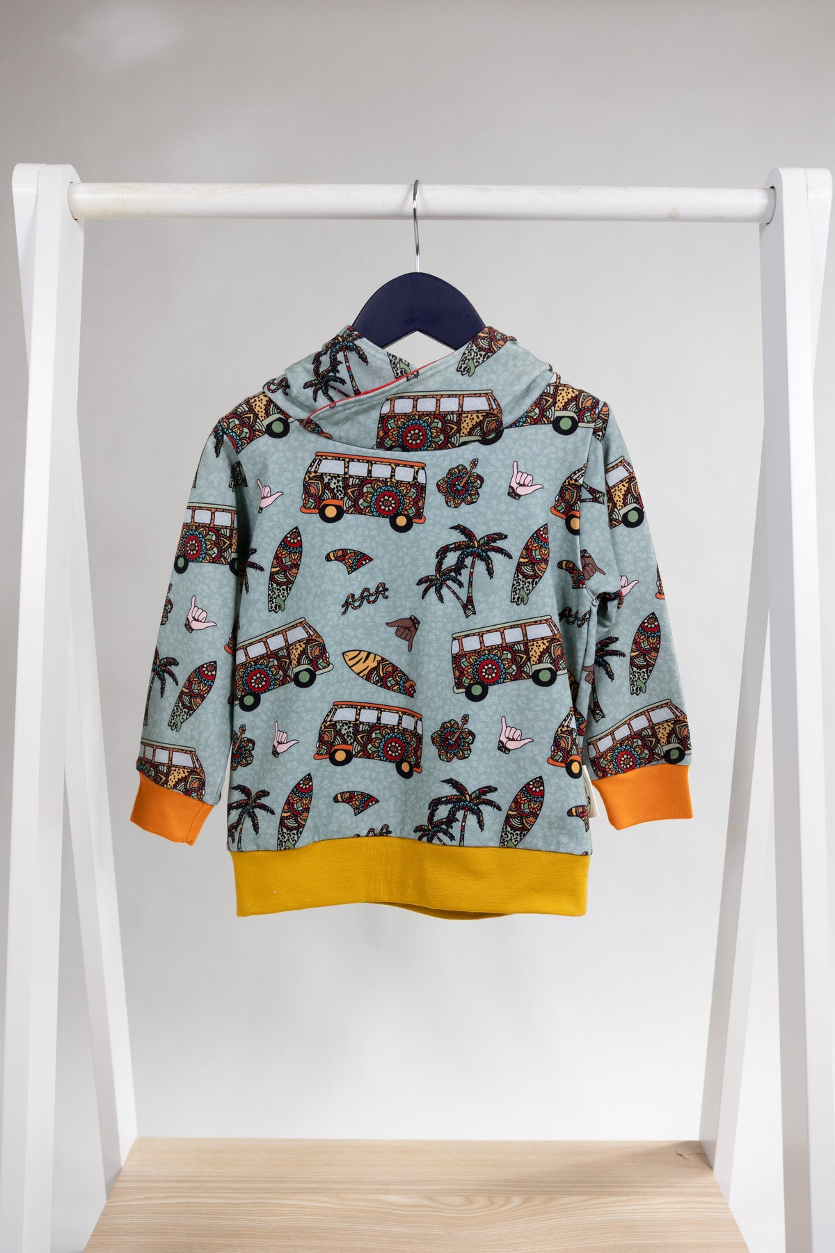 A children's hooded sweatshirt hanging on a black hanger from a white clothing rack. The sweatshirt has a light blue-gray background with a colorful print of vintage buses, palm trees, and surfboards, with bright orange cuffs and a yellow hem.