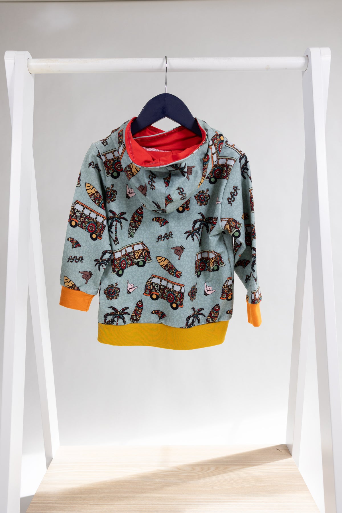 A children's long-sleeve hooded sweatshirt hanging on a black hanger from a white clothing rack. The sweatshirt has a light blue-gray base with a colorful print of red buses, palm trees, and surfboards, with a bright red inner hood lining and a mustard yellow hem and cuff trims.