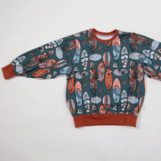 A long-sleeve crewneck sweatshirt featuring an all-over surfboard print in orange, blue, and white on a dark teal background, with solid rust-brown ribbed cuffs, collar, and hem.